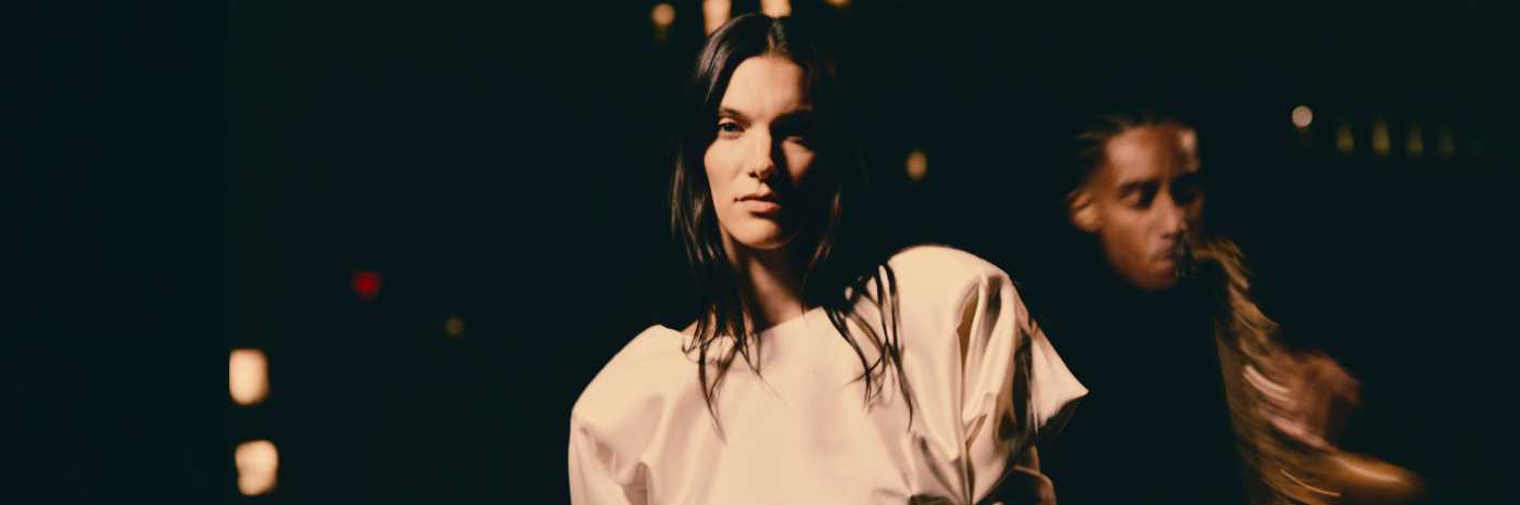 Close-up promotional image featuring Charlotte Cardin in a light-colored structured outfit illuminated by dramatic stage lighting. The person stands in sharp focus against a dark background, while a second blurred figure appears behind them. Warm highlights and deep shadows create a cinematic concert or runway atmosphere.