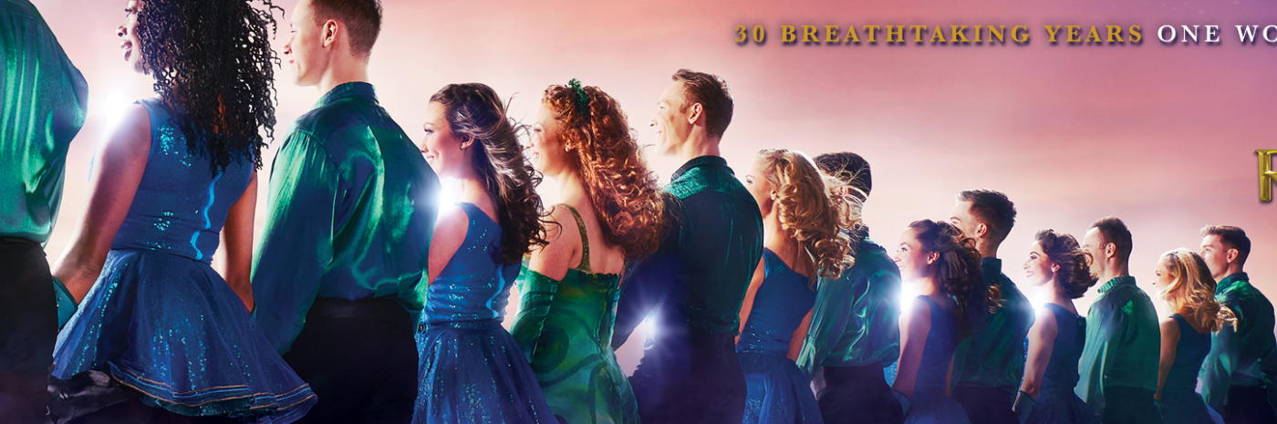 Promotional banner for Riverdance 30: The New Generation featuring a line of dancers in blue and green costumes illuminated by bright stage lighting against a pink and purple sky background. Gold text reads, “30 Breathtaking Years One Worldwide Phenomenon,” alongside the Riverdance 30: The New Generation logo. The dancers are shown in profile, standing shoulder to shoulder across the width of the image.