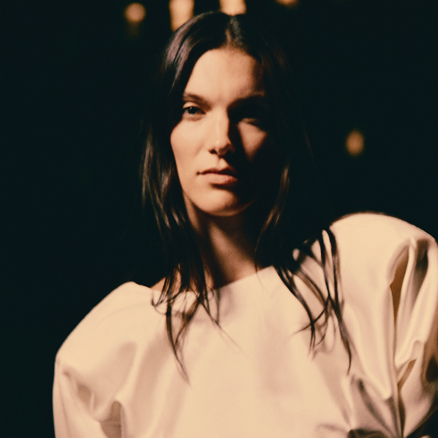Close-up promotional image featuring Charlotte Cardin in a light-colored structured outfit illuminated by dramatic stage lighting. The person stands in sharp focus against a dark background, while a second blurred figure appears behind them. Warm highlights and deep shadows create a cinematic concert or runway atmosphere.